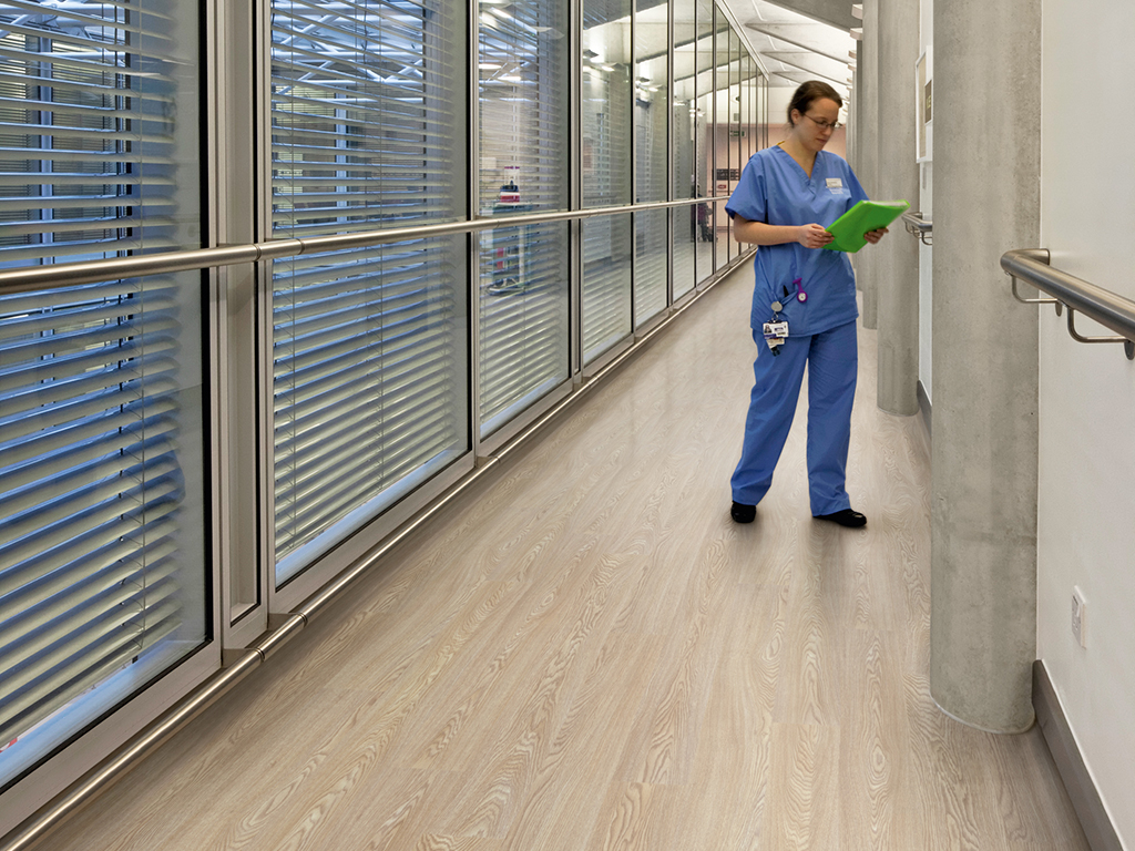 UCH Macmillan Cancer Centre, London, United Kingdom. Architect: Hopkins Architects Partnership LLP, 2012. Glass and bare concrete corridor with nursing staff.
