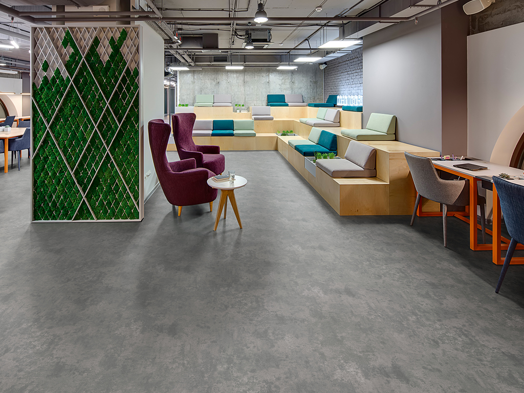 Interior in a loft style with a big zone with wooden benches and seats. Benches are decorated with grass, plants and bricks. On the right there is a gray table with orange legs and two chairs. In the 